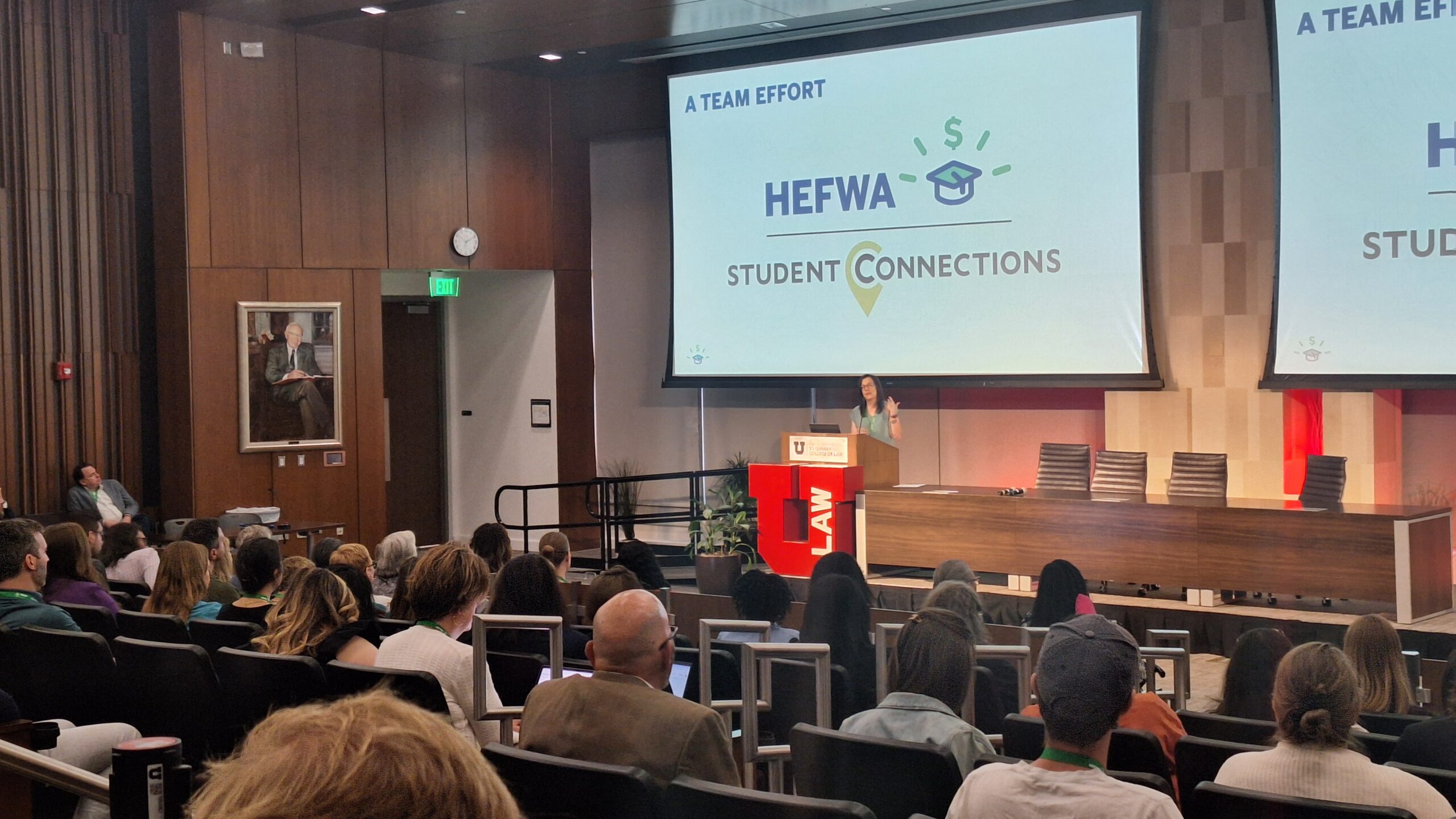 A medium-sized auditorium that’s three-quarters full. A woman is at the podium, behind her, two large screens display screenshots of the CashCourse application. An audience of about 200 individuals takes notes on the presentation.